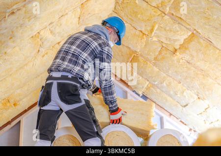 A construction worker in a blue helmet and checkered shirt is installing insulation in a home's attic during daylight. Safety gear is worn for protect Stock Photo