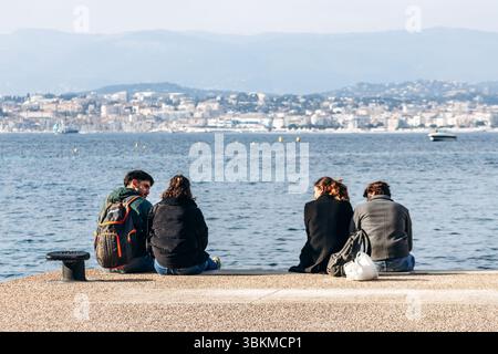 Cannes, France - March 8, 2025: Group of friends sitting by the sea on Sainte-Marguerite Island with Cannes city view. Stock Photo