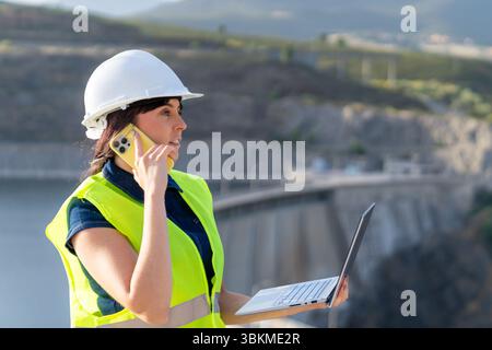Female engineer managing infrastructure, talking on phone and using laptop near dam Stock Photo