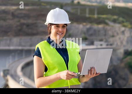 Female engineer using laptop managing infrastructure near dam Stock Photo