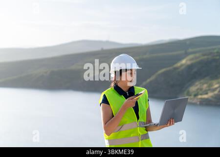 Female engineer managing water reservoir infrastructure using laptop and smartphone Stock Photo