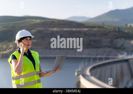Female engineer using laptop and talking on phone at dam Stock Photo