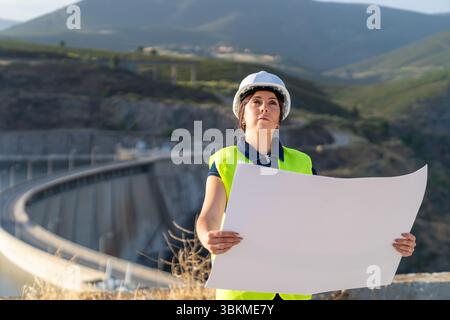 Female engineer inspecting dam construction with blueprint, standing in front of a mountainous landscape Stock Photo