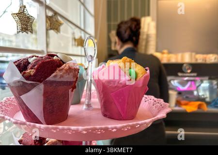 Close-up of colorful muffins and assorted baked goods arranged on a pink plate. Soft focus reveals a woman in the background, likely in a kitchen Stock Photo