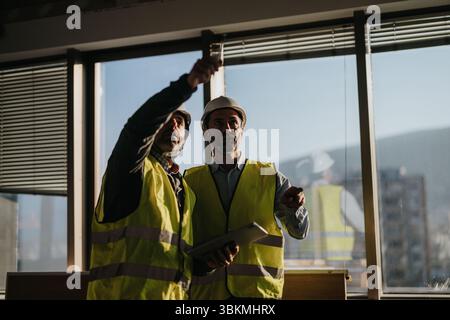 Construction professionals collaborating inside a building during daylight hours, inspecting architectural plans and discussing project progress. Stock Photo