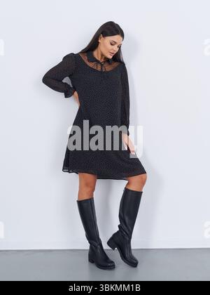 Dark-haired woman in knee-high black boots and sheer-sleeved dress poses against white wall, looking down. The dress has a delicate, intricate pattern Stock Photo