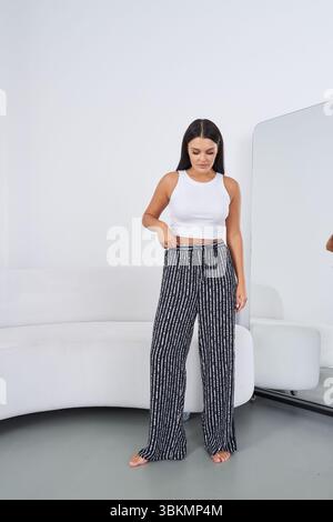 Young woman in a white sleeveless top and patterned black wide-leg pants stands barefoot near a curved white couch and large mirror on a light gray Stock Photo