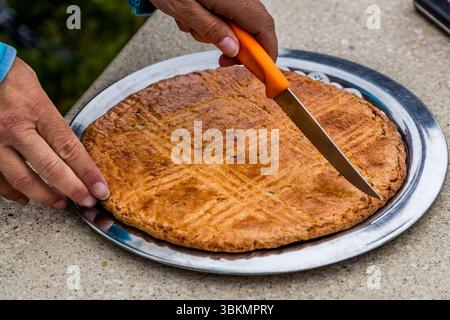 A "Galette Charentaise" is being cut. The cake speciality from the Charente contains candied stems of the medicinal plant angelica (Angelica archangelica), and the small green pieces are visible in the dough. The round galette lies on a metal plate, its surface scored with a diamond pattern. Tour guide Katell Nicoux cuts it with an orange-handled knife. The galette is part of the picnic on the guided cycling tour by Die Landpartie on the banks of the Charente at Le Ponton, Rouffiac, Nouvelle-Aquitaine, France. Stock Photo