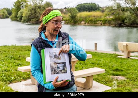 Tour guide Katell Nicoux from Die Landpartie explains that the nutria (Myocastor coypus) living by the river in the Charente are also used for culinary purposes. She points to a picture of a nutria and a jar of pâté de ragondin (nutria pâté). The tour group uses breaks such as the picnic on the banks of the Charente at Le Ponton, Rouffiac, Nouvelle-Aquitaine, France, to learn about special features along the river. One of them is the high population of invasive nutria. The inhabitants combat it by processing the animals into rillettes. The group is on a guided cycling tour on the La Flow Vélo  Stock Photo
