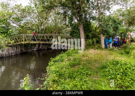 A narrow wooden bridge crosses the Charente at Les Gonds, Nouvelle-Aquitaine, France. Pedestrians and cyclists use the crossing. It is part of La Flow Vélo, a long-distance cycle route that runs for around 400 kilometres from Sarlat-la-Canéda in the Périgord to the Île d'Aix off the Atlantic coast. A person in a red jacket pushes a bicycle across the bridge. The rest of the group waits with helmets and e-bikes on the bank. The group is on a guided cycling tour by Die Landpartie along the Charente. Stock Photo