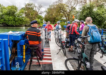 A ferryman in a striped jumper and a peaked cap sits on his stool at the blue drive unit, steering the Chaniers ferry (Bac de Chaniers) across the Charente. A group of cyclists, including journalist Angela Berg (back left, in a red jacket), crosses the river with their bicycles. The ferry takes them to the opposite bank of the Charente, from Chaniers, Nouvelle-Aquitaine, France, to Courcoury. The crossing dates back to the Middle Ages and is free of charge. The scene shows a relaxed atmosphere and the joy of cycling in nature. The group is on a guided cycling tour by Die Landpartie along the C Stock Photo