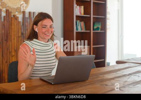 A woman wearing headphones smiles and waves while engaging in a video call. She sits at a wooden desk in a modern, well-lit workspace. Stock Photo