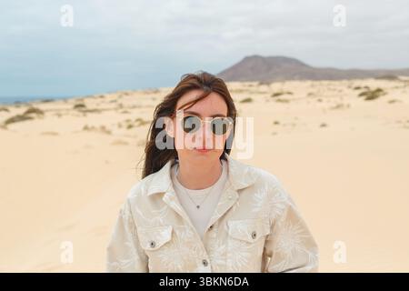 A woman stands in a vast desert, wearing sunglasses and a light jacket, gazing thoughtfully at the horizon with a mountain backdrop. Stock Photo