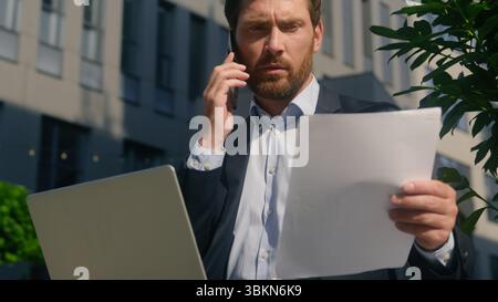 Serious business man mature middle-aged businessman multitask busy entrepreneur speaking smartphone holding documents in city laptop work report Stock Photo