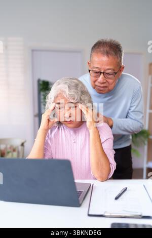 A concerned Asian older husband supports his stressed wife working on her laptop at home, offering comfort and care, reflecting elderly support and pr Stock Photo