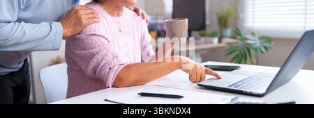 This image shows the hand of an Asian older woman using a laptop while holding coffee supported by her husband in a cozy home, capturing technology an Stock Photo