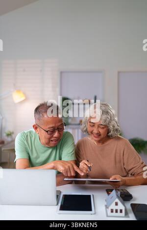 An older Asian couple happily reviews financial documents together at home using a laptop, tablet and calculator planning for future property investme Stock Photo
