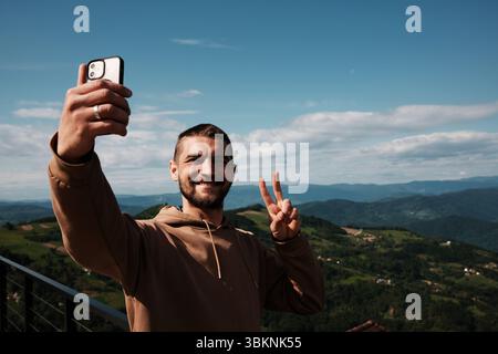 Cheerful young bearded man taking a selfie and showing a peace sign with Serbian mountains in the background. Male travel blogger Stock Photo