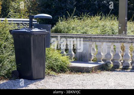 a black trash can with a lid and a small, attached ashtray or trash can standing on a dirt path next to a white stone balustrade and green bushes. Stock Photo