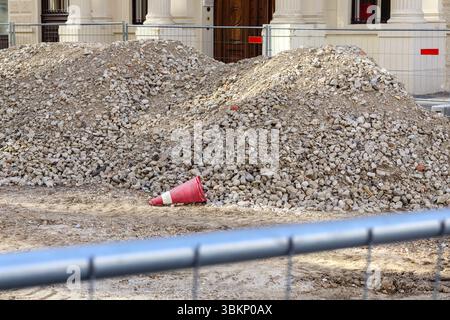 A scene of a construction site with two large piles of gravel or rubble, a red cone lying in the foreground, and part of a silver metal fence. Stock Photo