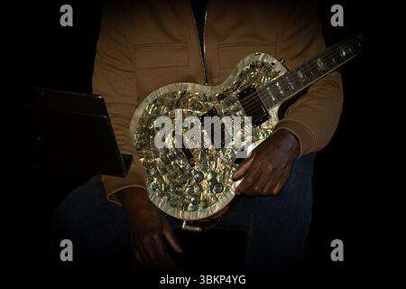 A man in a brown jacket is seated on stage holding a multi colored silver guitar. The guitar glows under a bright spotlight. Stock Photo