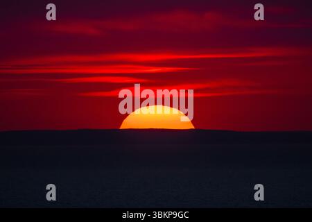 Vibrant red sunset over the sea horizon with the sun half-hidden behind a dark silhouette of distant hills. Stock Photo