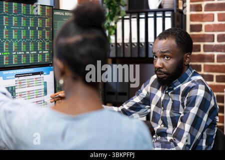 Selective focus on black male manager explaining financial charts on computer screen to female coworker. African american colleagues working together in analyzing forex trading data in modern office. Stock Photo