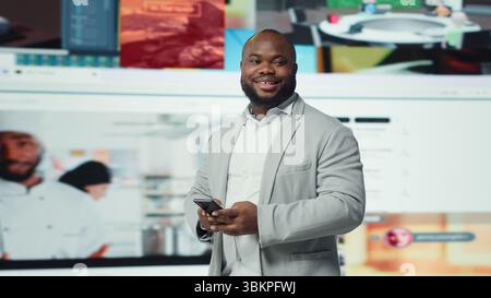 Portrait of smiling man browsing social networks feed on smartphone in front of videowall. Jolly african american person uses phone, floating icons highlighting interactive engagement online, camera A Stock Photo