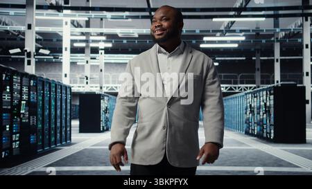 Smiling employee walking in high tech data center with machines powering networks, running companies databases. Admin overseeing server farm with rigs modules processing large datasets, camera A Stock Photo