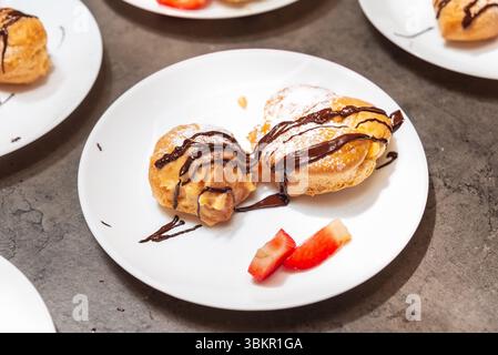 Two cream puffs topped with powdered sugar and chocolate sauce, served with fresh strawberry slices on a white plate. Stock Photo