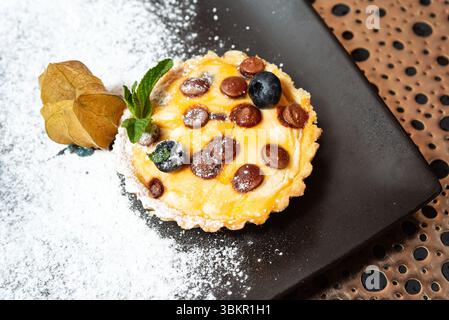Close-up of a mini tart topped with chocolate chips, blueberries, and mint leaves, dusted with powdered sugar on a dark plate. Stock Photo