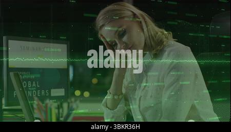 Reviewing data, woman in white blouse using laptop in office at night, with data overlay Stock Photo