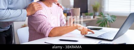 This image shows the hand of an Asian older woman using a laptop while holding coffee supported by her husband in a cozy home, capturing technology an Stock Photo