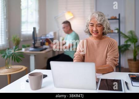This image shows a senior Asian woman using a laptop at home. Her husband is working nearby. It shows the online business lifestyle of older people wi Stock Photo