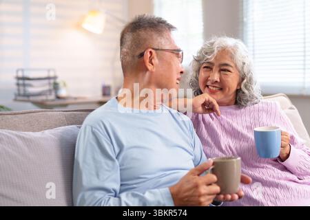 A delightful photo of an older Asian couple enjoying a quiet moment at home. They are smiling, drinking coffee and spending their retirement years tog Stock Photo