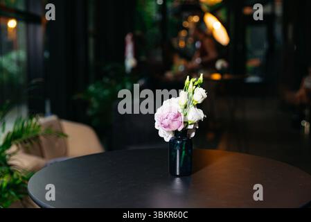 A delicate bouquet of white and purple flowers in a dark vase sits on a round table in a cozy, dimly lit café. Stock Photo