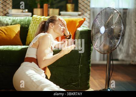 An adult woman sits on the floor, hand to her forehead with eyes closed, visibly suffering from the intense summer heat despite a nearby standing fan, Stock Photo