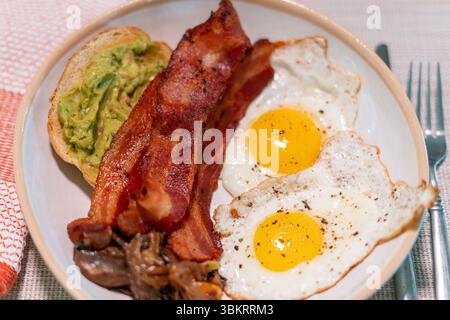 Hearty breakfast plate with crispy bacon, fried egg, sautéed mushrooms, and creamy avocado toast on rustic ceramic dish—perfect for brunch menus. Stock Photo