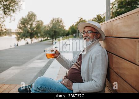 Senior man enjoying beer on sunny riverside promenade Stock Photo