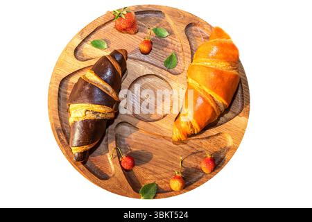 Top view of classic and chocolate croissants with wild strawberries and leaves on a round wooden tray. Isolated food composition for bakery menu or de Stock Photo