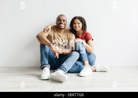 Happy young black couple sitting on floor, leaning against white blank wall Stock Photo