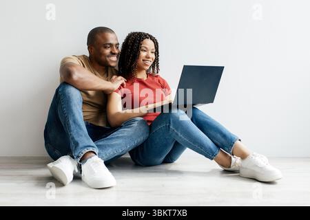 Young Black couple sitting on floor at home, smiling while using laptop together Stock Photo