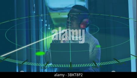 Wearing red headphones, IT technician holding open laptop in server rack aisle, with AR graphics Stock Photo