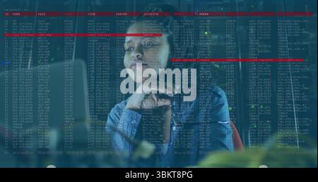 Mid adult blue-shirted woman studying code on glass panel at office, with monitors and keyboard Stock Photo