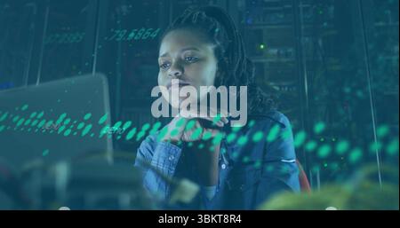Monitoring woman wearing denim shirt analyzing system on laptop in server room, with blinking racks Stock Photo