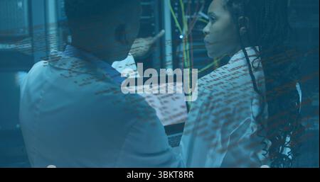 Pointing techs in lab coats examining server racks and Ethernet cables in server room, with laptop Stock Photo