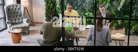 Website header shot of diverse group of young adult and middle aged professionals collaborating around table in modern office with plant wall, while using digital tablet and working with documents Stock Photo