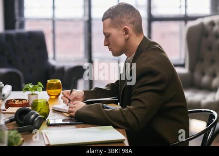 Caucasian young adult man sitting at desk writing in notebook during business meeting in modern office workspace, focused on taking notes with documents and headphones nearby Stock Photo