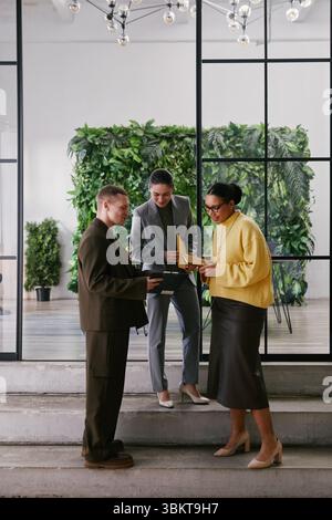 Vertical shot of two young adult women and man standing together in modern office discussing business documents, collaborating on project near glass wall with greenery Stock Photo