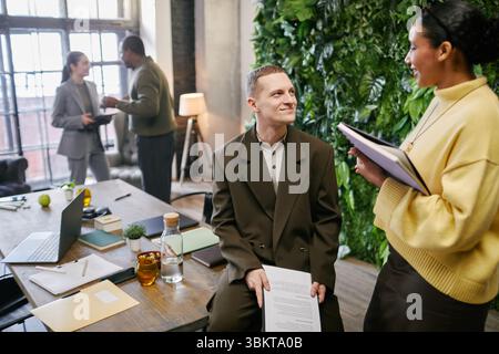 Caucasian young adult man sitting on desk holding documents smiling at Black young adult woman standing with folders, while multiethnic colleagues discussing in background in office Stock Photo